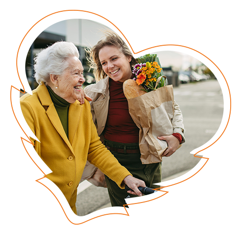 The image shows two individuals, an older woman and a younger person, standing together on a street, with the older woman holding a bouquet of flowers and both smiling. They appear to be engaged in a pleasant interaction, possibly sharing a moment of connection or celebration.