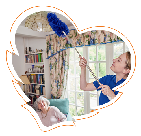 The image shows a woman cleaning a ceiling fan with a duster while standing next to an elderly person seated on a couch, both indoors with a heart-shaped design overlaying the scene.