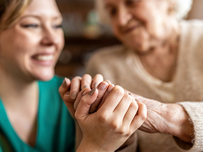 The image shows two individuals, likely an older adult and a younger person, holding hands with smiles on their faces, suggesting a moment of affection and connection between them.