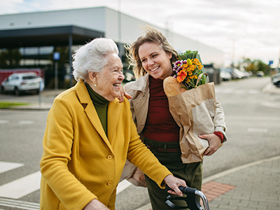 An elderly woman pushing a cart with flowers on a street corner, accompanied by a younger woman who appears to be assisting her.