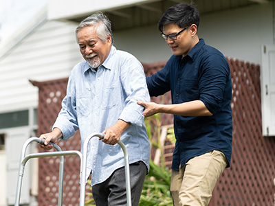 The image shows an elderly man using a walker being assisted by a younger person who appears to be pushing him forward.