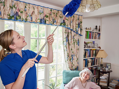 Woman in blue uniform holding a duster cleaning a ceiling fan.