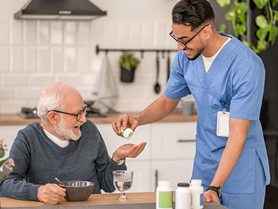 The image depicts an elderly man receiving assistance from a healthcare professional at his home, with various medical items visible on a dining table between them.