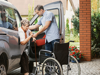 A man in a blue shirt is assisting an elderly woman with a walker into a car.