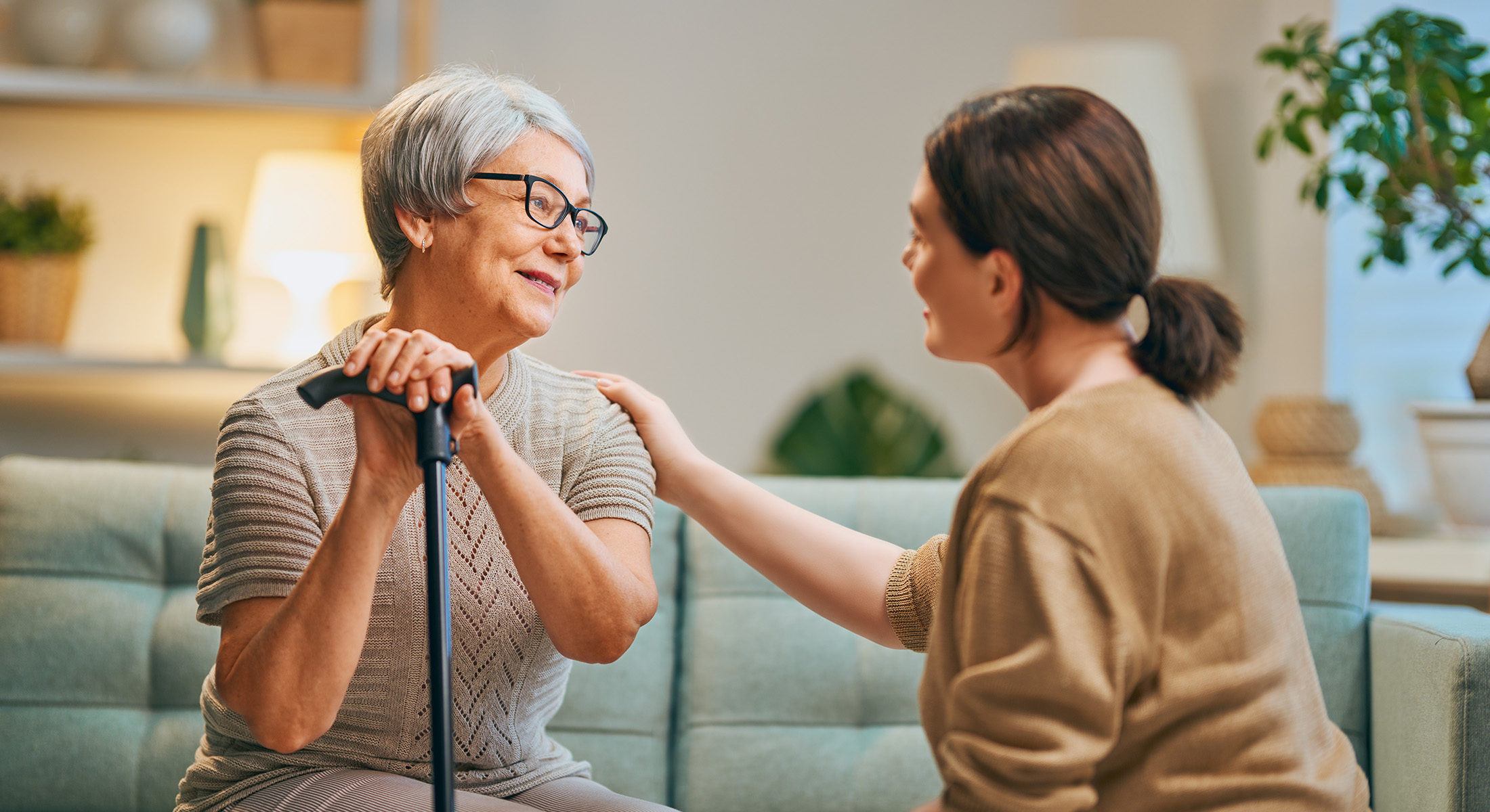 The image shows two elderly individuals sitting on a couch with one person holding a cane, engaging in an interaction that appears to be supportive or comforting.