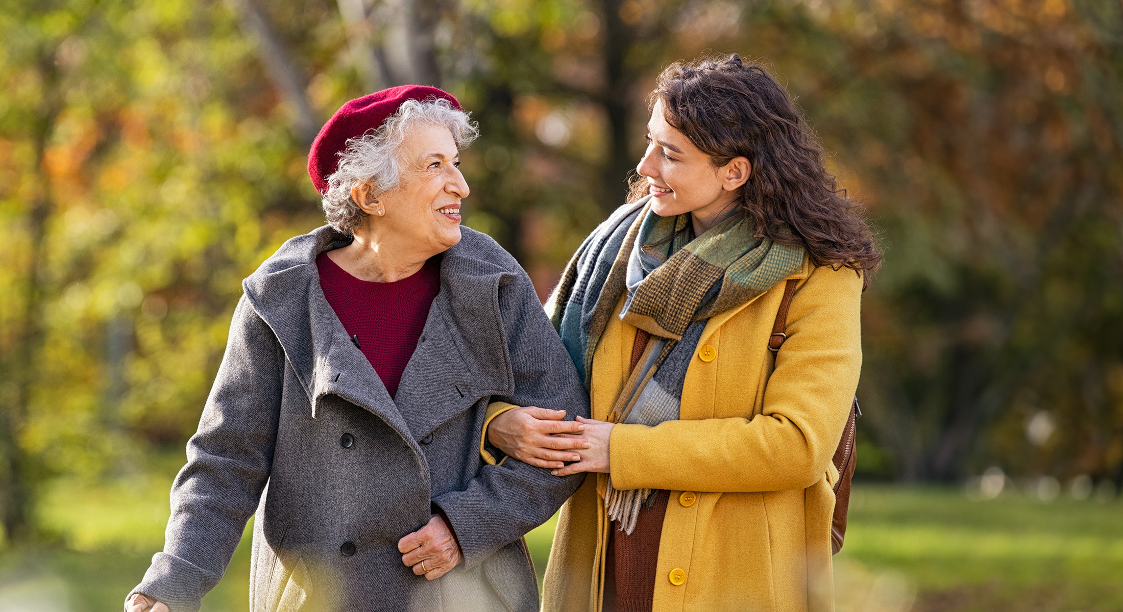 The image shows two individuals walking side by side; an older woman with a cane and a younger person, both smiling and engaged in conversation, set against a park-like background during autumn.