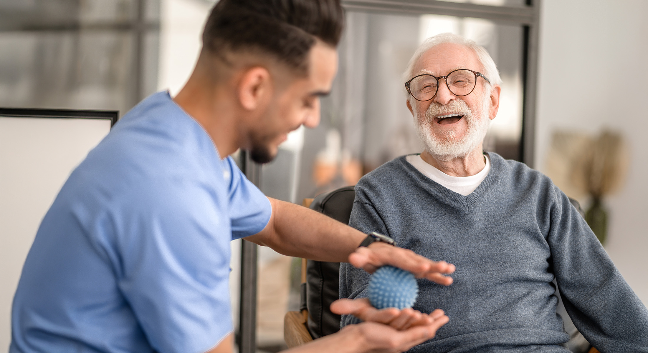 A young man in scrubs is assisting an elderly gentleman with a massage, both are seated in a modern living room.